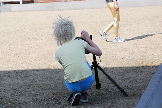 A man wearing blue tights, a yellow shirt and a blonde wig is crouched over his camera. Of in this the distance you see the subject he is photographing, her legs in golden pants.
