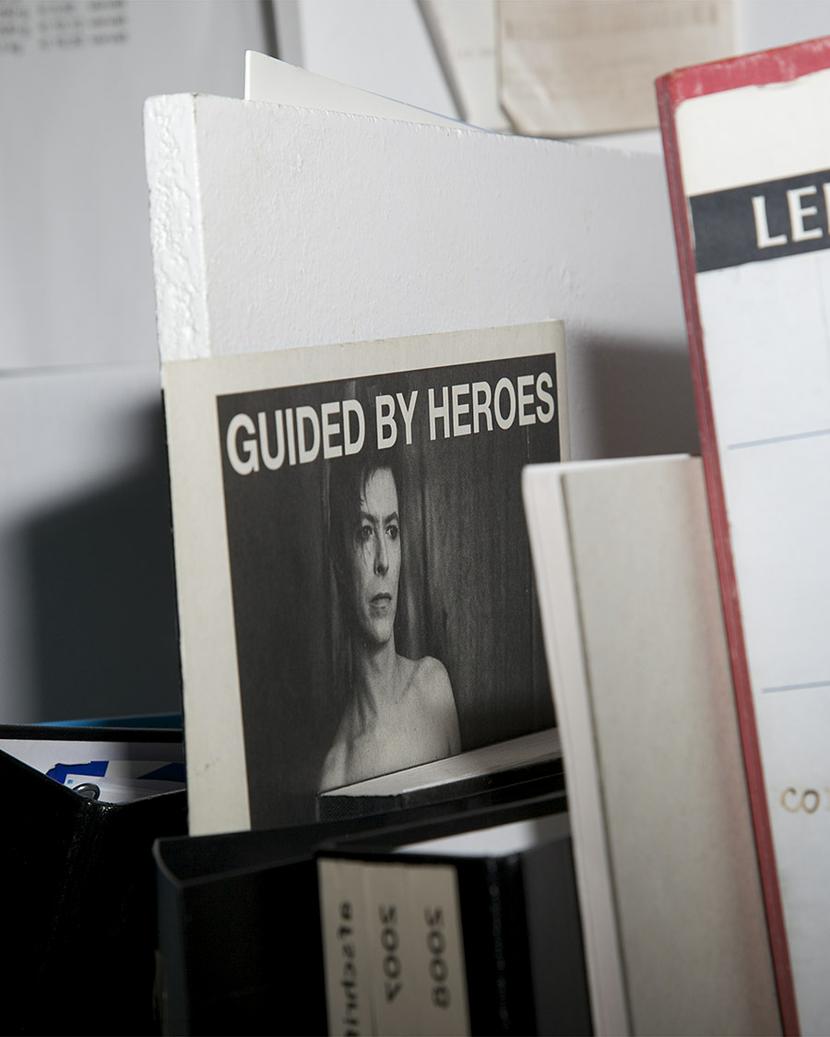 You see a picture of a small poster stuck behind some files. It says 'Guided by Heroes' and it shows David Bowie.