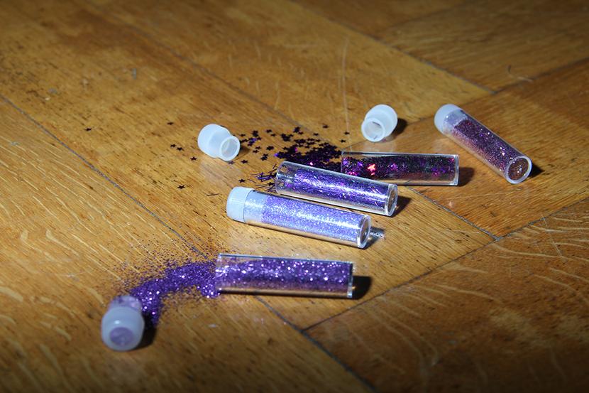You see different kinds of purple glitter emerge from little tube shaped containers spread out over herringbone parquet. The picture is lit by a flash
