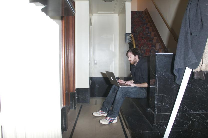 You see a young man sitting at the bottom of a staircase. The staircase ends in a marble piece and the floor is marble as well. The young man looks into the screen of his laptop.