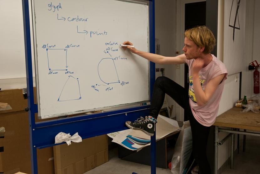 You see a young man posing to the right of a whiteboard, looking into the camera. He holds a marker in his right hand as if he is writing, his right foot is planted on the whiteboards horizontal support. He wears black leggings and a salmon pink sleeveless t-shirt. On the whiteboard is constructed a path of curves and lines, showing also the bézier control points. All the points are marked either offCurve or onCurve.