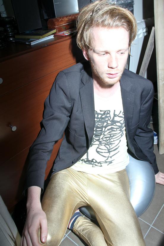 You see a young man perched against a dresser, sitting on a medicine ball. He looks away. He is wearing a slate blazer, over a yellow shirt, screenprinted with a drawing of his guts and his chestcage. He wears gold colored leggings. His hair is combed over to the left.