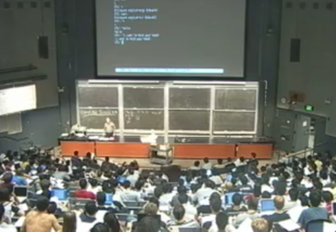 You see a lecture hall from above. Computer code is projected above the blackboards that contain formulas. The professor stands behind his desk. Many students have computers. To the left one large student wears no shirt.