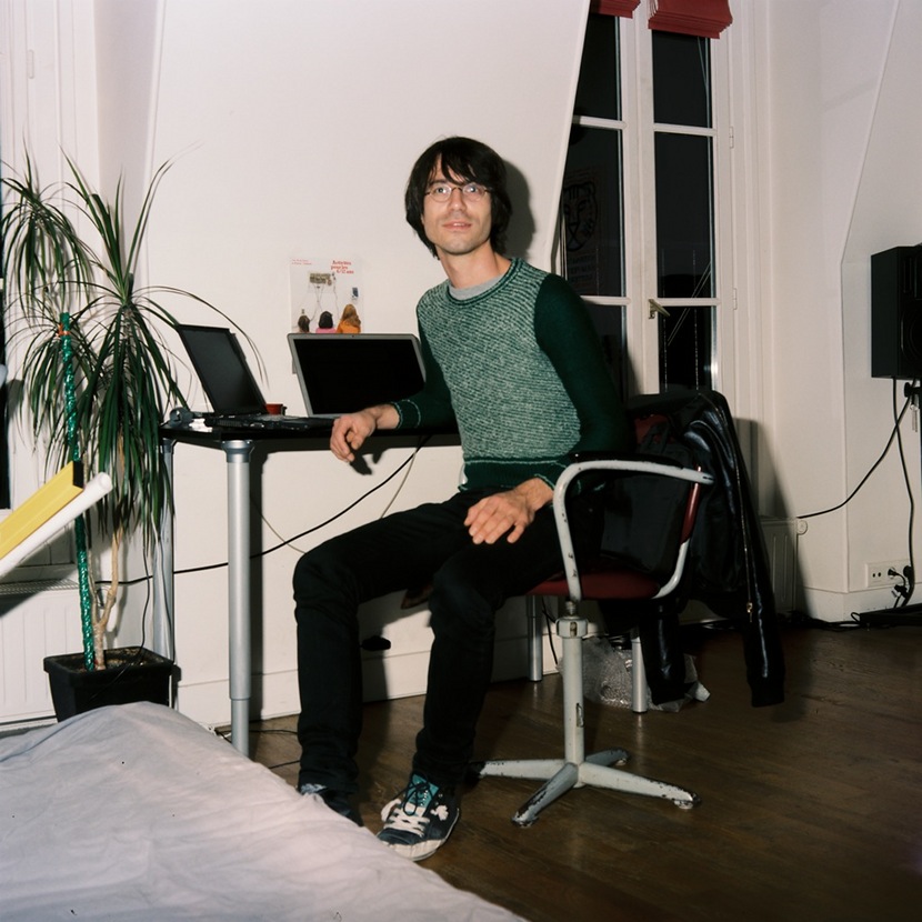 You see a young man looking at the camera. He sits on a chair in an appartment.